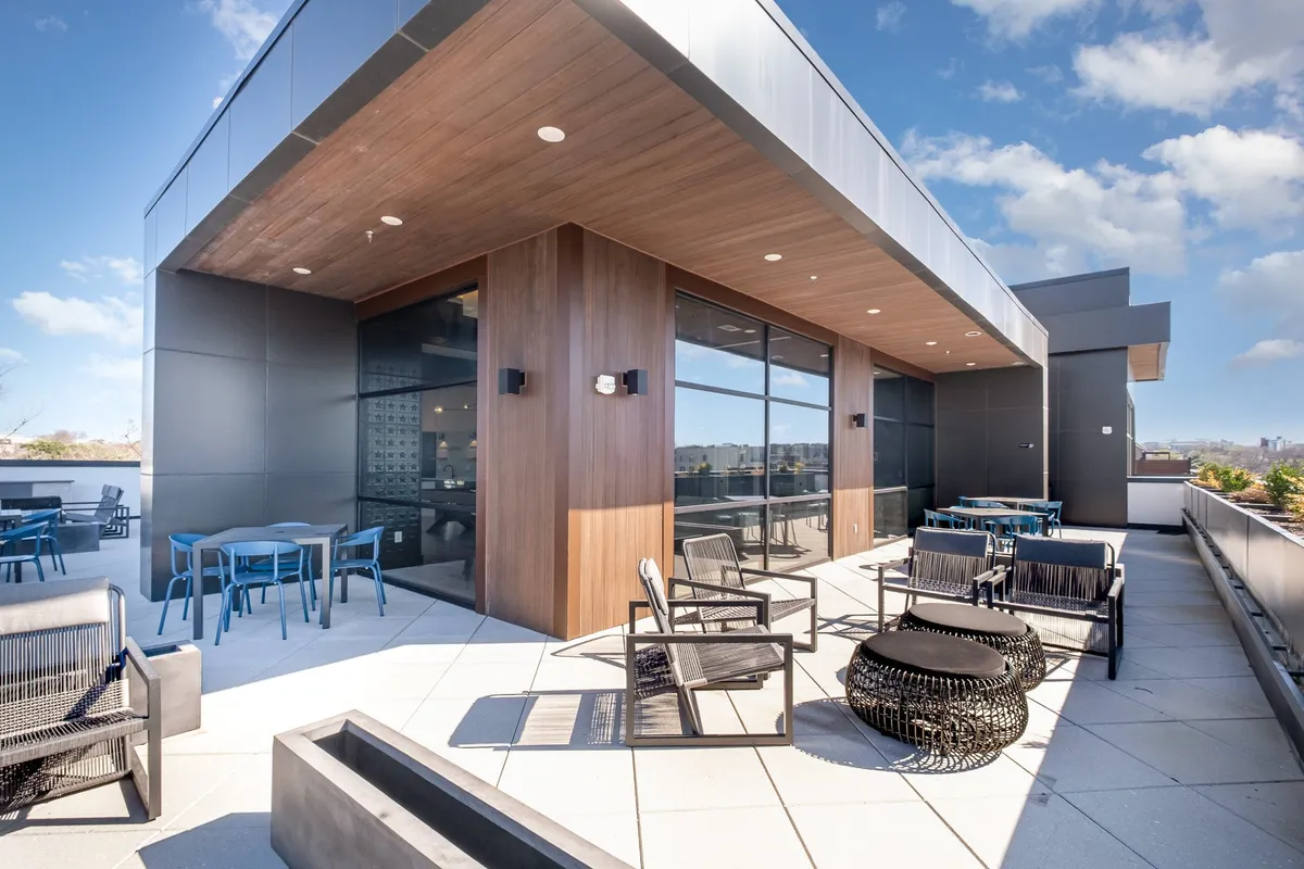 A contemporary outdoor lounge space featuring modern furniture, large windows, and a stylish wooden ceiling. The area is designed for social gatherings, relaxation, or al fresco dining, with natural light filtering in and a view of the city surroundings.