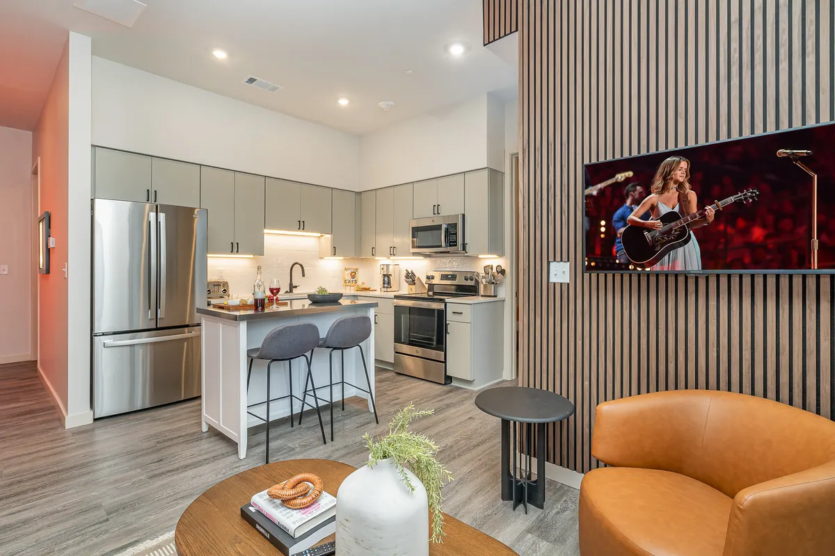 A modern interior combining the kitchen and living spaces, featuring stylish appliances, a seating area with an armchair, and a TV playing a live performance. The space feels lively and comfortable, ideal for both entertainment and relaxation.