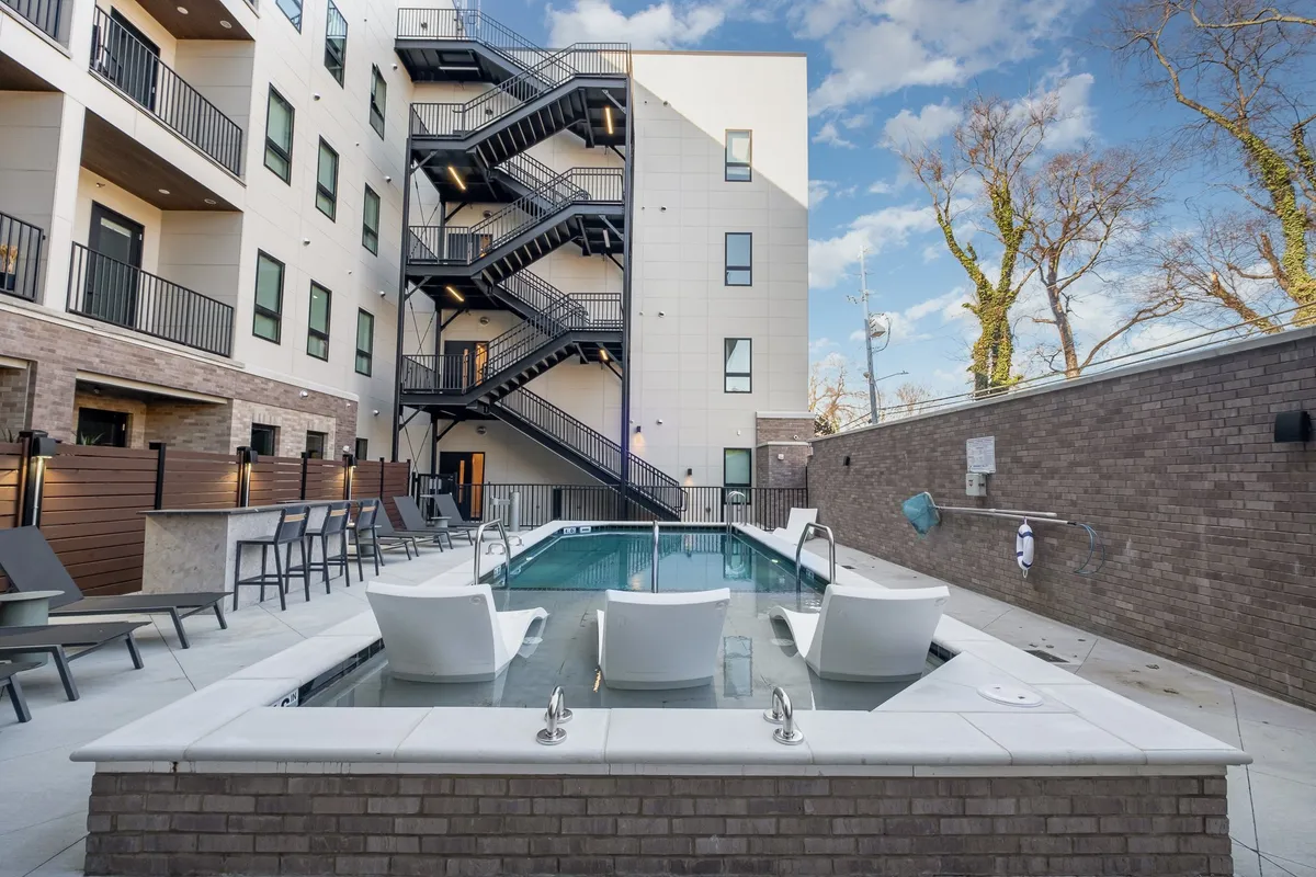 A beautifully maintained swimming pool surrounded by lounge chairs and a modern building facade, with a staircase leading up from the poolside. The area basks in natural light, creating an inviting environment for swimming, sunbathing, and gathering with friends or family.