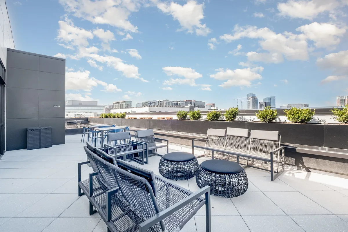 A stylish rooftop lounge with modern seating, lush planters, and a sweeping view of the city skyline at dusk. The setting is relaxed and inviting, perfect for unwinding with friends or enjoying a romantic evening under the stars.