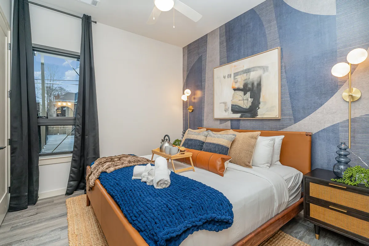 A modern and cozy bedroom featuring a bed draped with a blue blanket, abstract wall art above the headboard, and a nightstand adorned with a small plant. Natural light filters into the room, highlighting the comfortable and stylish atmosphere.